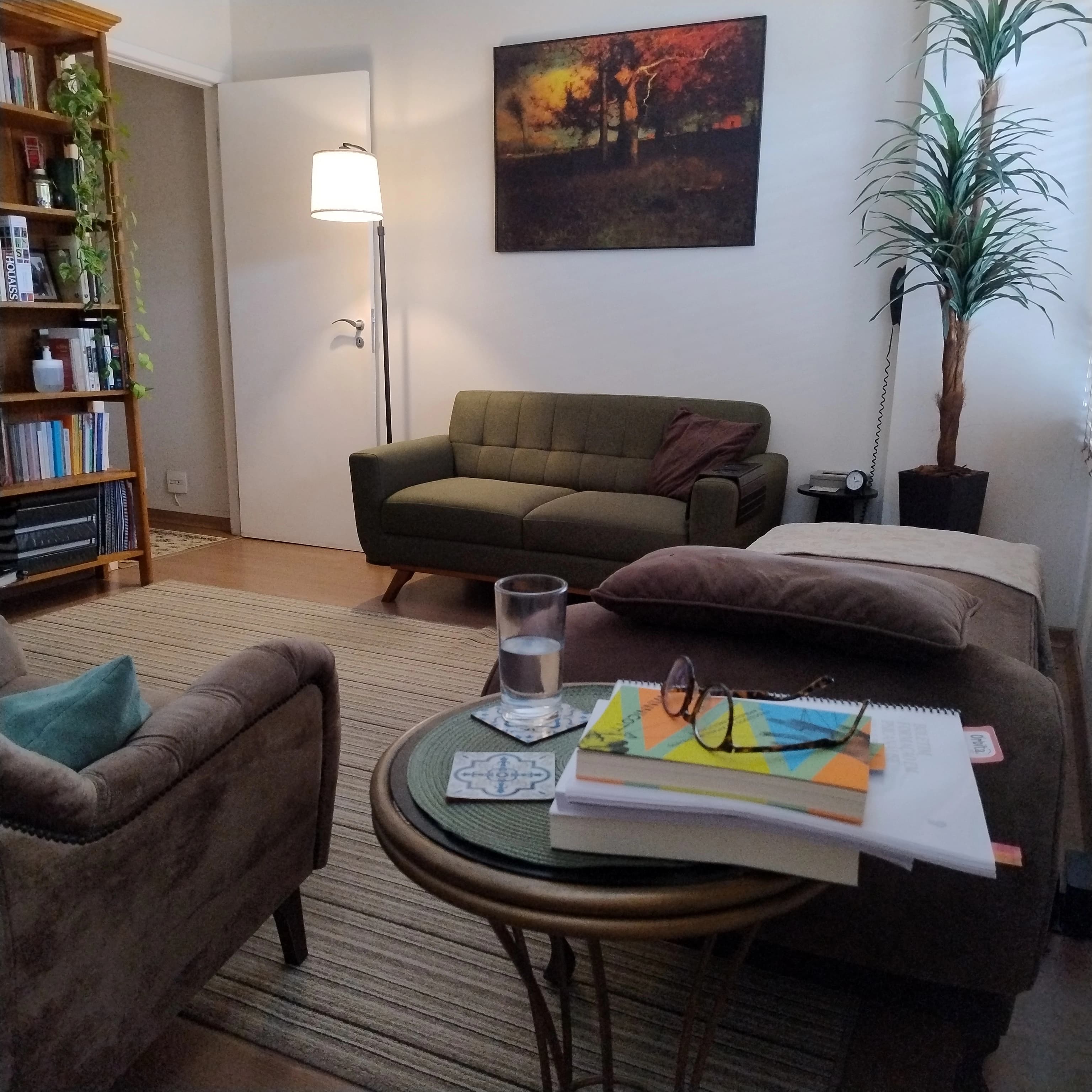 Consultório onde os atendimentos acontecem. Sala branca, com três sofás marrons, uma mesa com livros em cima, e uma estante cheia de livros.