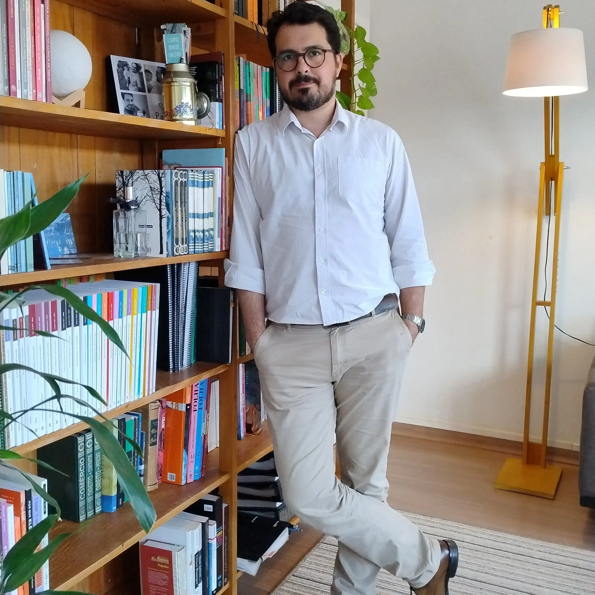 Psicólogo Helder em pé, encostado na estante de livros de seu consultório, vestindo uma camisa de botão branca com mangas longas, calças beges e sapato marrom. As mãos dele estão nos bolsos da calça e a manga longa está dobrada até o cotovelo. Possui um relógio prata em seu pulso.
